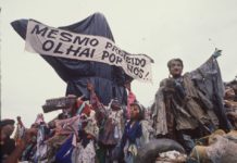 The photo shows the sculpture "Cristo Mendigo" that was the opening car of the Ratos e Urubus parade. In the image, the sculpture is covered in garbage bags as a censure, and carries the banner that reads "Even forbidden, watch over us!"