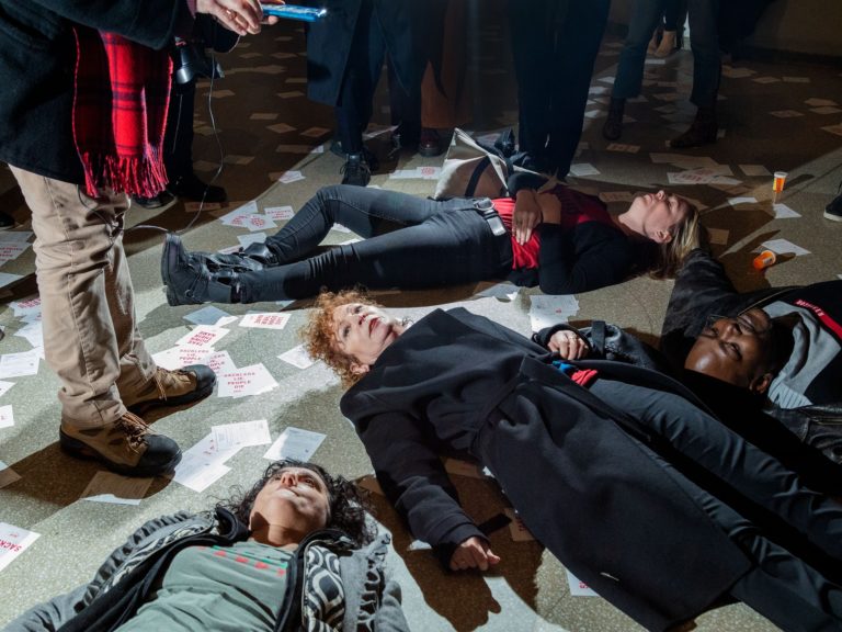 The photo shows Protestants performing a "Die in". The demonstration is against the Sackler family, linked to Purdue Pharma, producer of Oxy Contin. Among the protesters is photographer Nan Goldin, surrounded by papers that say "Sackler Lie People Die" and bottles of medicine.