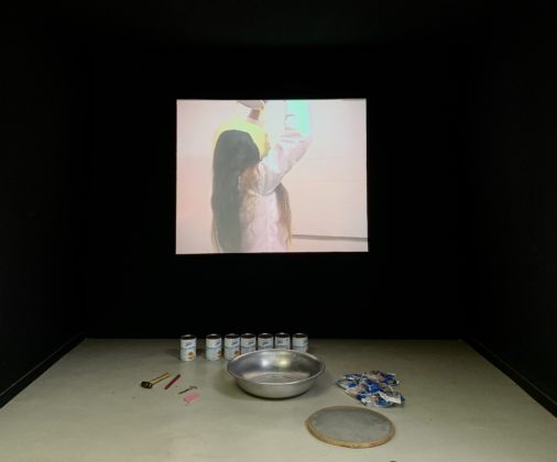 Exhibition piece Ratos e Urubus - The photo shows the exhibition of a recording of the performance "Pancake" by Márcia X. Below the screen where the video is projected are objects equivalent to those used by her in her performance.