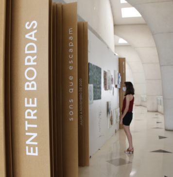 No primeiro plano, divisões de madeira na parede tem o escrito "Entre Bordas", na vertical. Ao fundo, uma mulher olha quadros da exposição "Entre bordas - sons que escapam" na parede