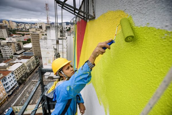 A artista Criola, sobre um andaime, pintando a empena do edifício Chiquito Lopes com um tom de amarelo
