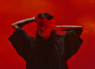 A performer Emilie Sugai está no centro da foto, vendando seus próprios olhos com uma faixa vermelha. A iluminação da imagem é toda vermelha, contextualizando um dos momentos do espetáuculo AKA