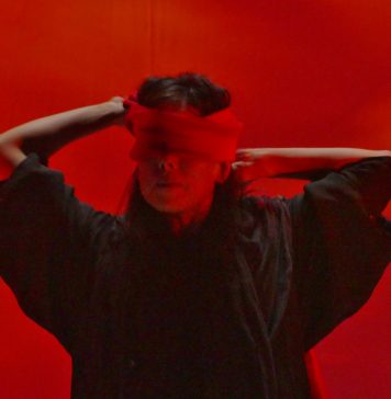 A performer Emilie Sugai está no centro da foto, vendando seus próprios olhos com uma faixa vermelha. A iluminação da imagem é toda vermelha, contextualizando um dos momentos do espetáuculo AKA