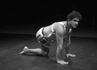 Horizontal photo, black and white. In the midst of the choreography, João Paulo Lima has both hands and the only knee on the floor, keeping his back aligned, on a plank over his knee. He's in profile. He uses a costume that refers to the practices of bondage and sadomasochism, with most of the skin exposed, semi-nude. This photo is a still of the show DEVOTEES, presented in the program Zona de Criação, from the Cultural Hub of Ceará PORTO DRAGÃO.