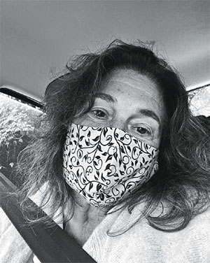 Vertical photo, black and white. Patrícia Rousseaux, Editorial Director of artebrasileiros, appears in the foreground. She wears a white mask with the silhouette of leaves and branches as a pattern. Her eyes have a calm expression. She has shoulder-length wavy hair and her head slightly tilted to the left.