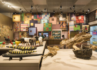 Horizontal, color photo. View of the MIRROR COUNTRIES exhibition. In the foreground, sculptures of animals, some colored, in wood, others in straw. In the background, a mural with capulanas and fabrics from different regions of Angola, Mozambique, Guinea Bissau, São Tomé and Principe, Cape Verde and Brazil. Kazuo Kajihara's photo.