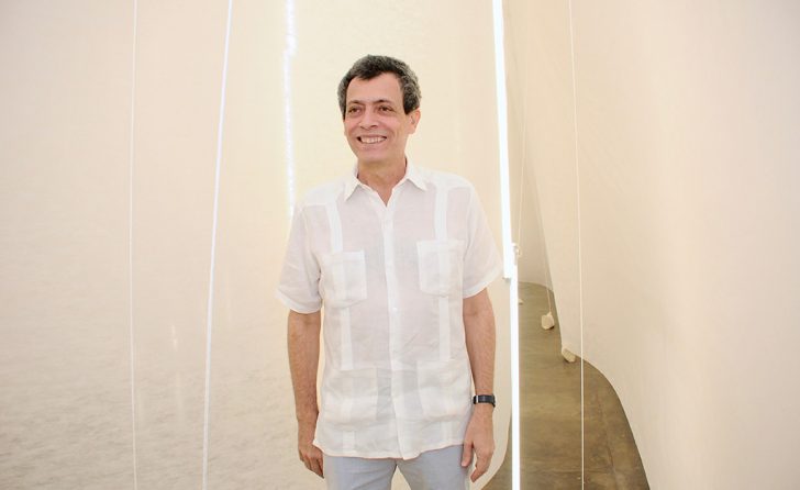 Foto horizontal, colorida. Carlito Carvalhosa está de pé, usa uma camisa branca de mangas curtas e uma calça cinza claro. Sorri. Ao fundo, a Instalação FAÇO TUDO PARA NÃO FAZER NADA.