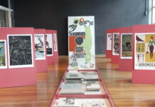 Horizontal, color photo. View of the ILLUSTRATED WORKERS exhibition, curated by Chico Homem de Melo. On the left and right sides, red panels bear prints of worker illustrations. In the background, in the center, the expansion, by about two and a half meters, of the FABRIL WORKER illustration by Odileia Toscano. In front of him, a long table, which takes the photographer to the panel, brings the original books and magazines, from which the illustrations were taken.