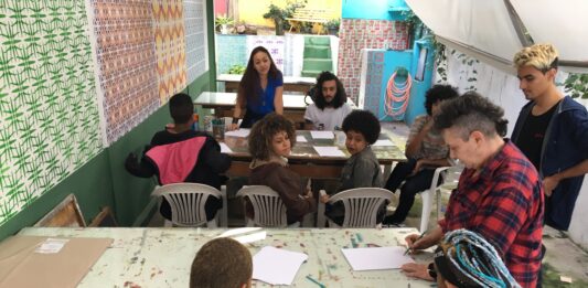 Imagem horizontal colorida. Um coletivo de artistas sentados em torno de duas mesas no ateliê do JAMAC conversam. Algumas folhas de papel sobre as mesas.