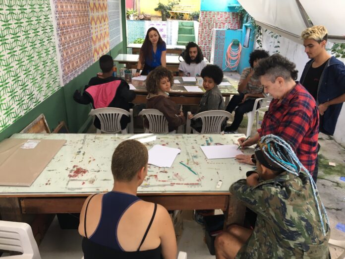 Imagem horizontal colorida. Um coletivo de artistas sentados em torno de duas mesas no ateliê do JAMAC conversam. Algumas folhas de papel sobre as mesas.