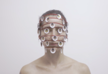 Horizontal, color photo. Work THOUSAND EYE, by Lia Chaia. In the background, a white wall. A white woman, with her eyes closed and her brown hair tied back. The photograph frames her from the shoulders up. She has bare shoulders. Seven red strands wrap its head horizontally and spaced apart. The first at the hairline, the others parallel, crossing the rest of the face, the last being at the height of your chin. Hanging on the wires, several round pieces refer to eyes, they are white with a brown circle inside and a black circle inside it, as if they were iris and pupil. The work is part of the exhibition SAY NO