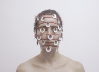 Horizontal, color photo. Work THOUSAND EYE, by Lia Chaia. In the background, a white wall. A white woman, with her eyes closed and her brown hair tied back. The photograph frames her from the shoulders up. She has bare shoulders. Seven red strands wrap its head horizontally and spaced apart. The first at the hairline, the others parallel, crossing the rest of the face, the last being at the height of your chin. Hanging on the wires, several round pieces refer to eyes, they are white with a brown circle inside and a black circle inside it, as if they were iris and pupil. The work is part of the exhibition SAY NO