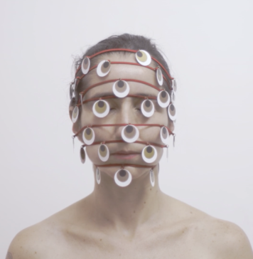 Foto horizontal, colorida. Obra MIL OLHOS, de Lia Chaia. Ao fundo, uma parede branca. Uma mulher branca, com os olhos fechados e os cabelos castanhos presos. A fotografia a enquadra dos ombros para cima. Tem os ombros nus. Sete fios vermelhos envolvem sua cabeça horizontalmente e de forma espaçada. O primeiro na linha do cabelo, os demais paralelos, cruzando o restante do rosto, sendo o último na altura de seu queixo. Penduradas nos fios, diversas peças redondas remetem a olhos, são brancas com um círculo castanho dentro e um círculo preto dentro deste, como se fossem íris e pupila. A obra é parte da exposição DIZER NÃO