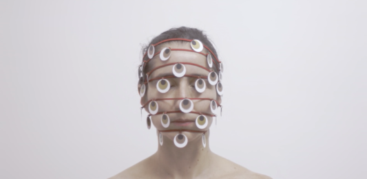 Horizontal, color photo. Work THOUSAND EYE, by Lia Chaia. In the background, a white wall. A white woman, with her eyes closed and her brown hair tied back. The photograph frames her from the shoulders up. She has bare shoulders. Seven red strands wrap its head horizontally and spaced apart. The first at the hairline, the others parallel, crossing the rest of the face, the last being at the height of your chin. Hanging on the wires, several round pieces refer to eyes, they are white with a brown circle inside and a black circle inside it, as if they were iris and pupil. The work is part of the exhibition SAY NO