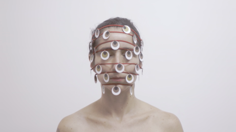 Horizontal, color photo. Work THOUSAND EYE, by Lia Chaia. In the background, a white wall. A white woman, with her eyes closed and her brown hair tied back. The photograph frames her from the shoulders up. She has bare shoulders. Seven red strands wrap its head horizontally and spaced apart. The first at the hairline, the others parallel, crossing the rest of the face, the last being at the height of your chin. Hanging on the wires, several round pieces refer to eyes, they are white with a brown circle inside and a black circle inside it, as if they were iris and pupil. The work is part of the exhibition SAY NO