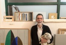 Foto horizontal, colorida. Julio Villani veste uma camisa branca e um casaco cinza. Está em seu ateliê, ao seu redor algumas obras e um papagaio.
