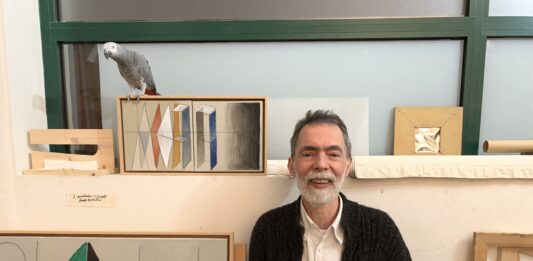Foto horizontal, colorida. Julio Villani veste uma camisa branca e um casaco cinza. Está em seu ateliê, ao seu redor algumas obras e um papagaio.
