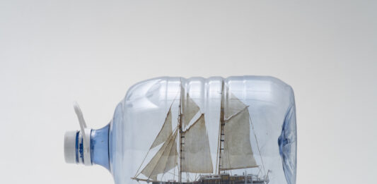 Imagem horizontal, colorida. Obra DRIFT BOTTLE 7 (TESTIGO DE DERIVA), de Jorge Macchi, uma garrafa plástica de água mineral que contém em seu interior a reprodução em escala reduzida de um veleiro